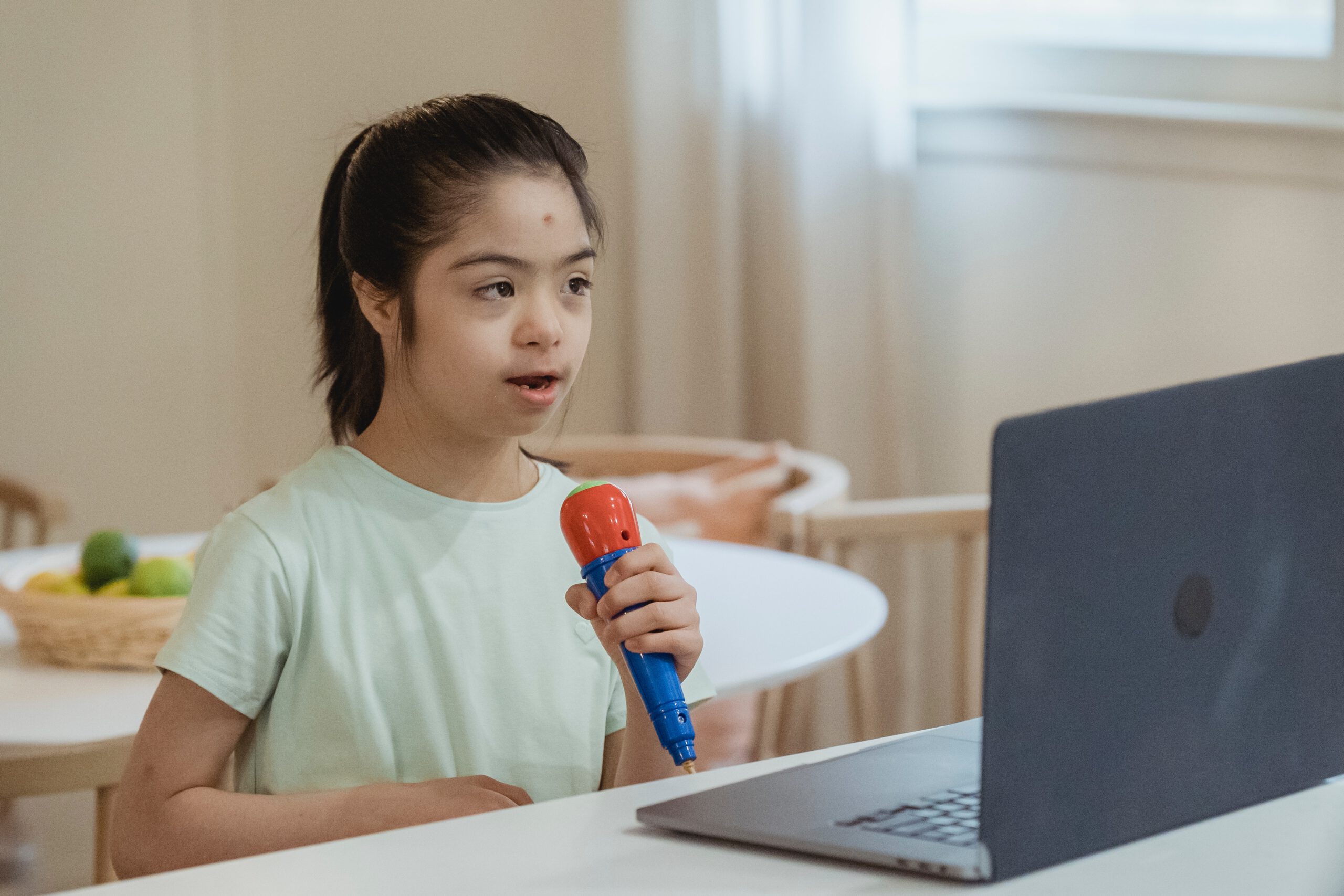 Dziewczynka z upośledzeniem umysłowym siedząca przed laptopem i trzymająca w ręce plastikowy zabawkowy mikrofon