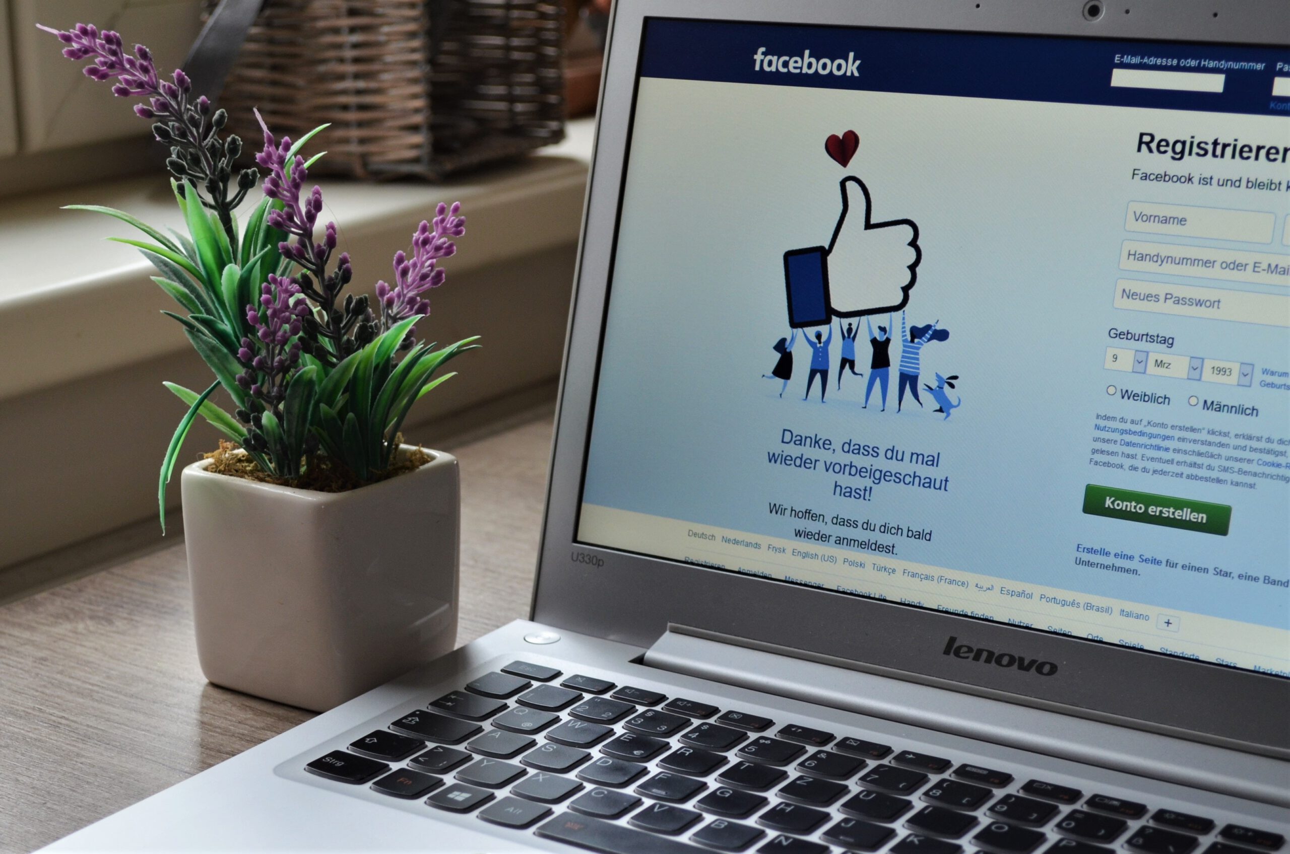 Otwarty laptop z wyświetloną stroną Facebooka na biurku i kwiatek w doniczce, stojący obok