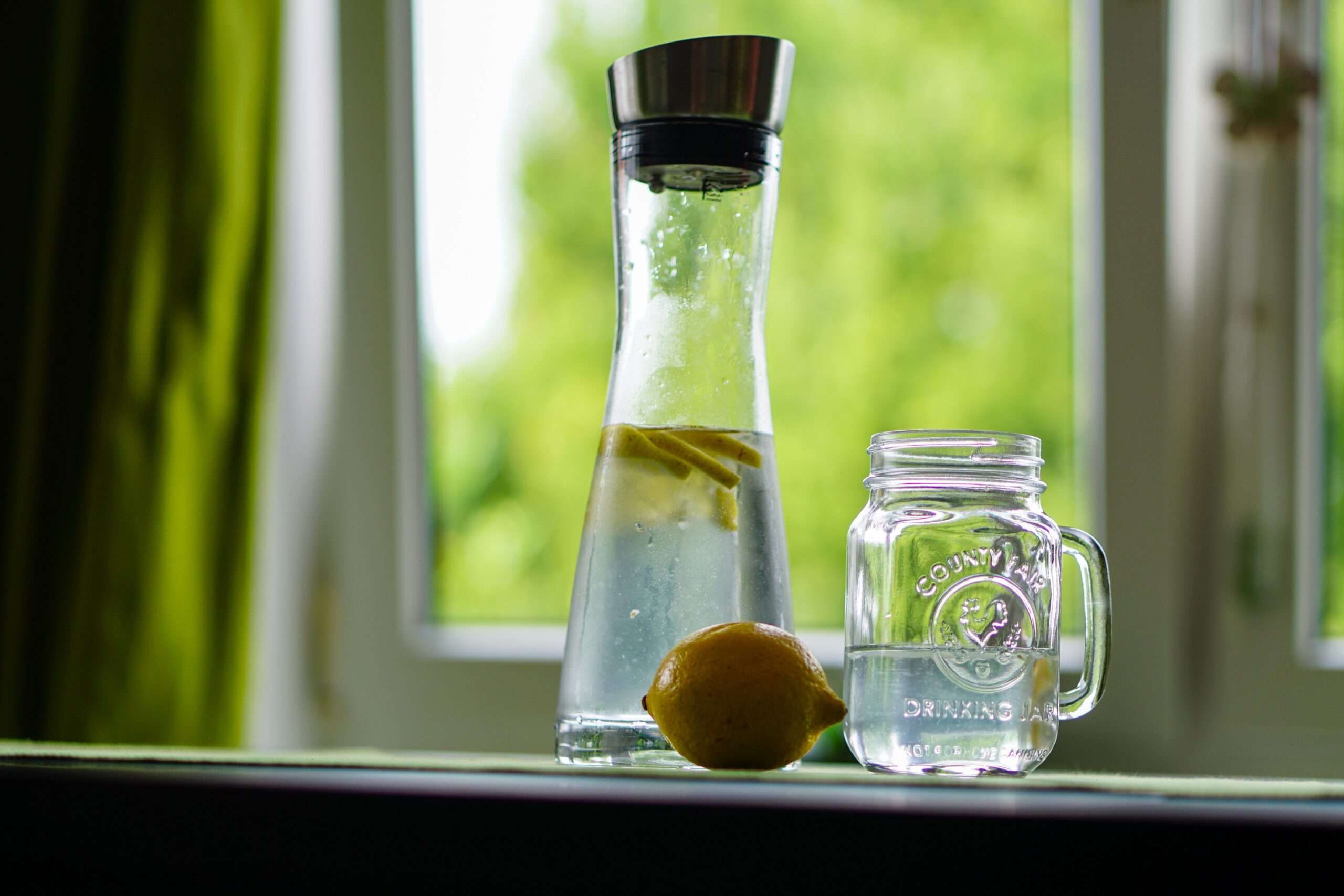 Szklana karafka z wodą, szklany kubek wypełniony do połowy wodą i cytryna na blacie na tle okna